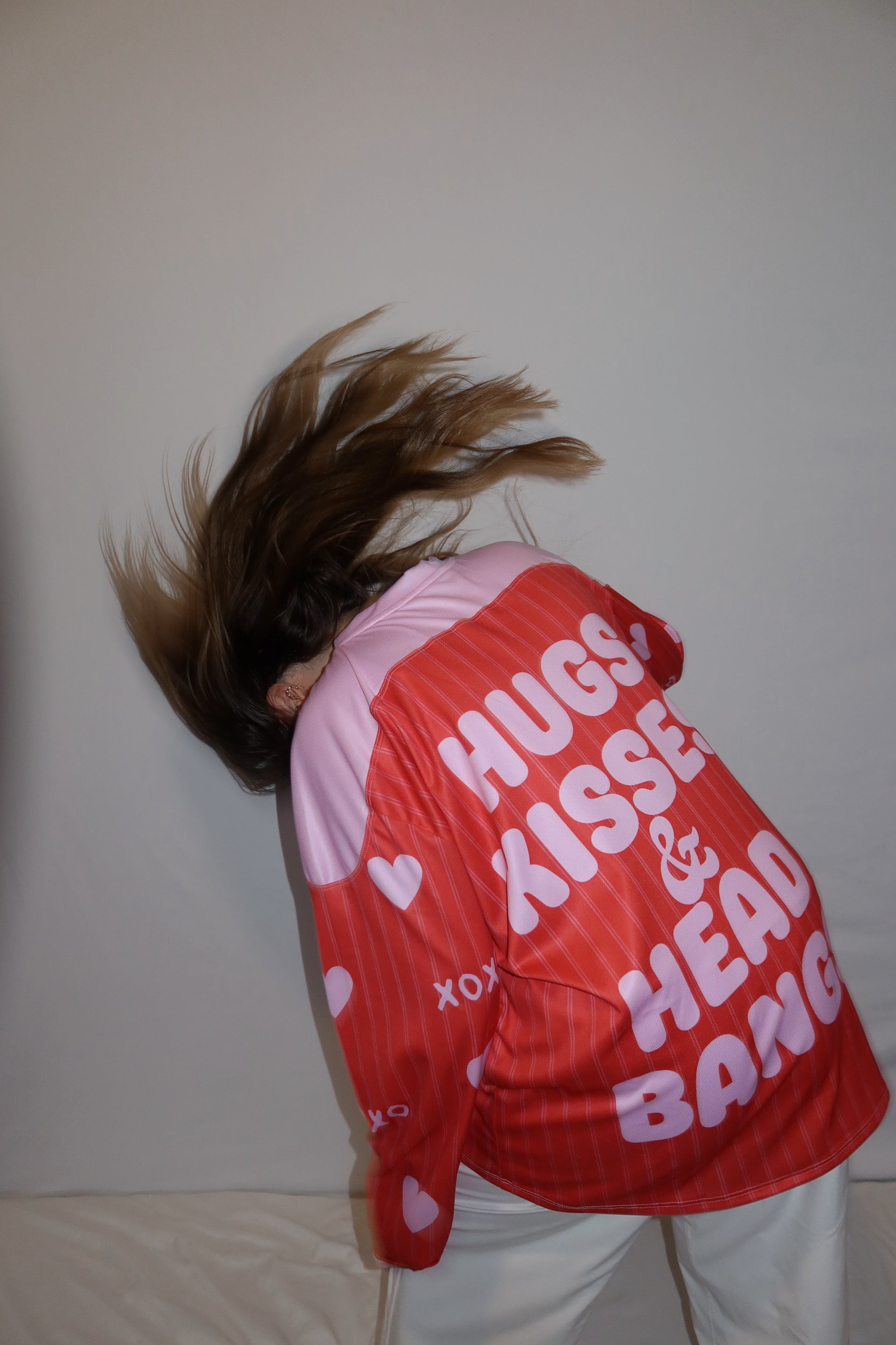Person head banging showing the back of a red and pink rave hockey jersey. The back says hugs, kisses, and head bangs. 